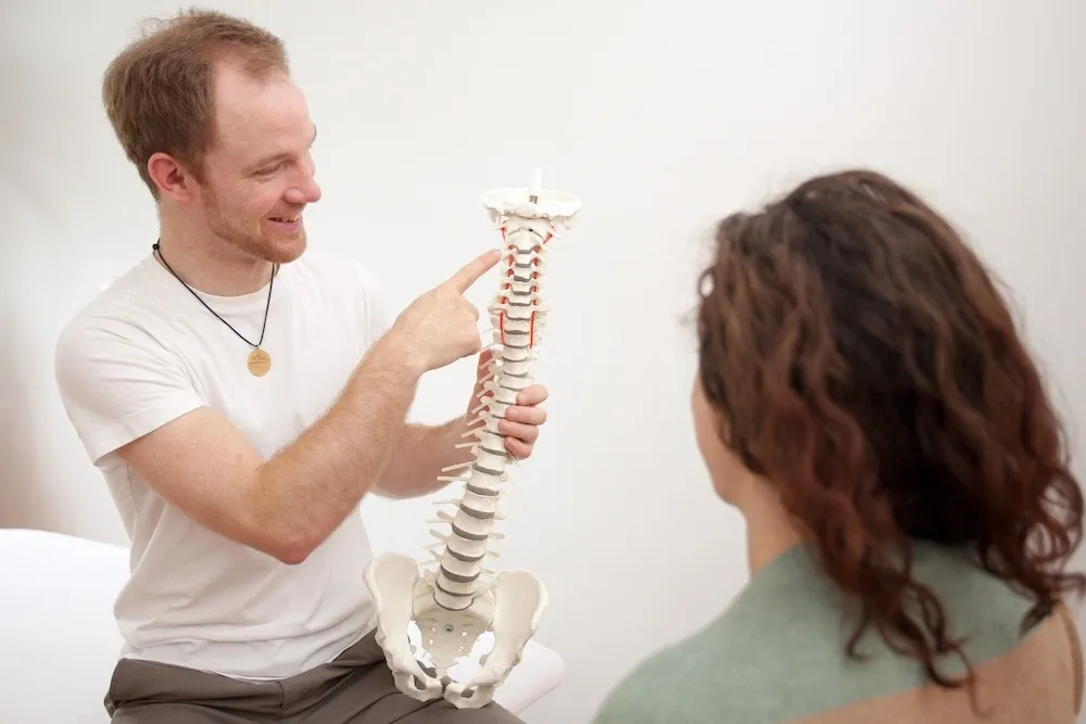 Osteopath Richard Manske erklärt ein Modell der Wirbelsäule. Eine Frau sitzt ihm gegenüber und hört ihm zu.